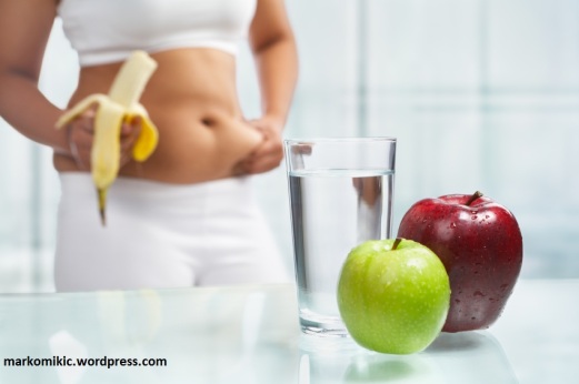 Apples and water on table with female body on sport attire eating banana at background, shot for diet concept
